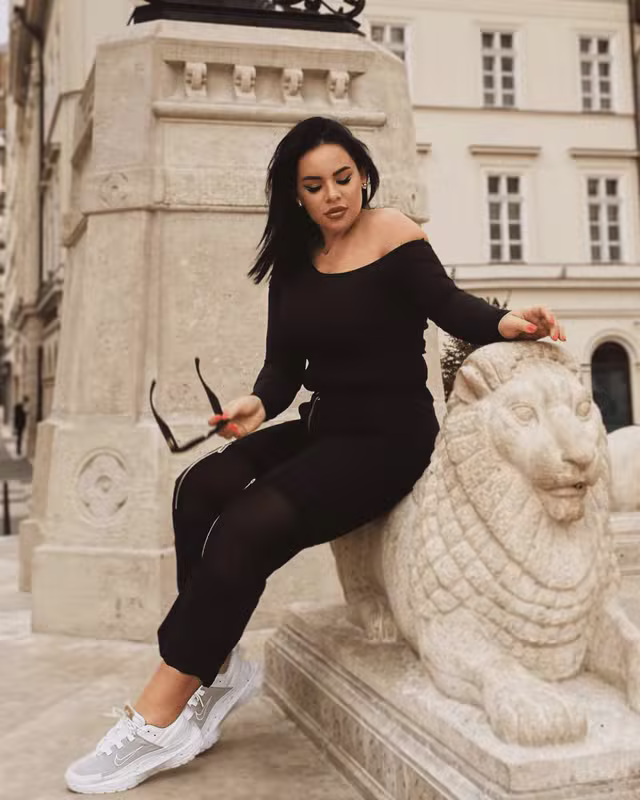 szidi.lila.free poses elegantly in an off-shoulder black outfit beside the stone lion.